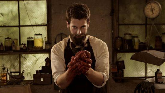 Director and writer Ryan Spindell poses in costume as a butcher holding a bloody heart in his hands.