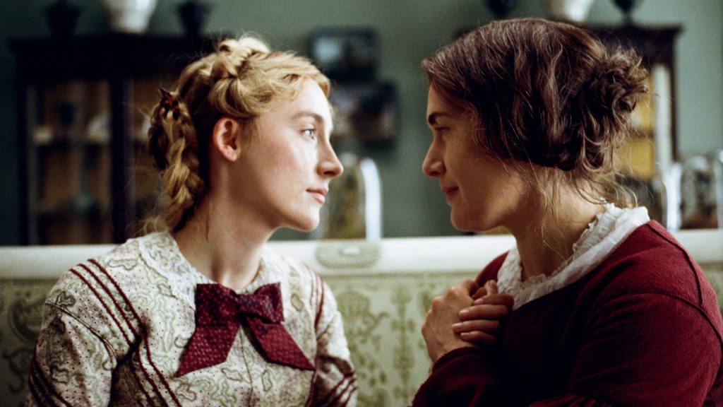 Kate Winslet and Saoirse Ronan hold hands together closely while both wearing passionate red clothing as seen in Ammonite.