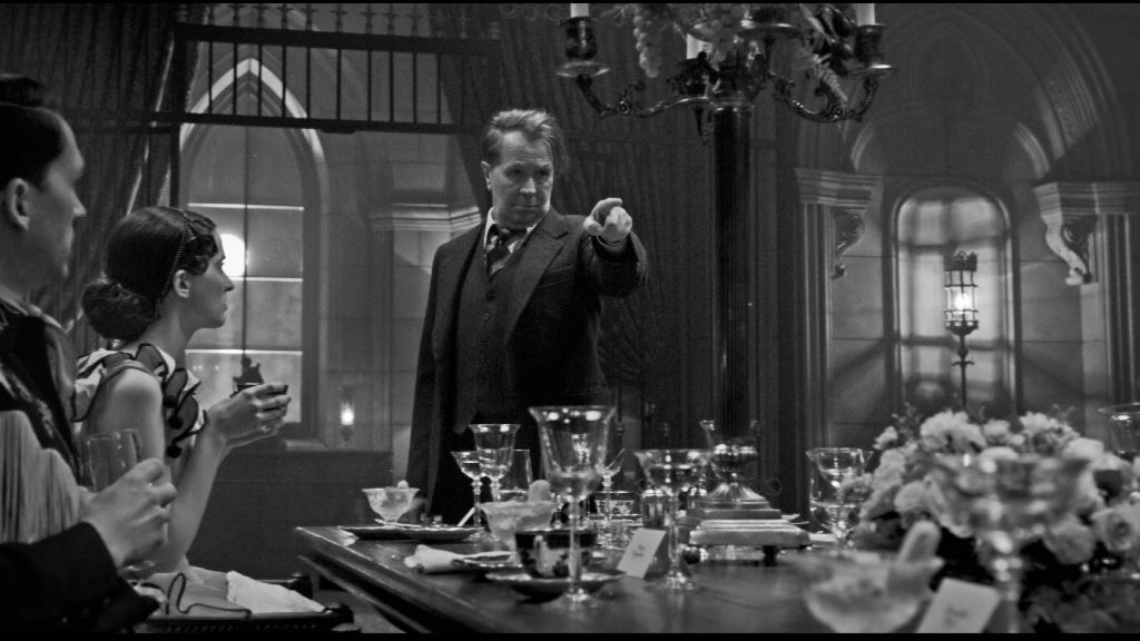 Gary Oldman as Herman J. Mankiewicz pointing his finger at a grand dinner table as seen in Mank. 