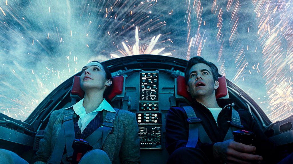 Gal Gadot and Chris Pine sit in the cockpit of a jet fighter as colorful fireworks blast around them from the outside as seen in Wonder Woman 1984.