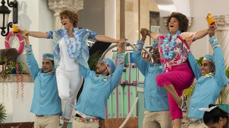Kristen Wiig and Annie Mumolo are lifted by hotel staff during a musical number inside a colorful resort as seen in Barb and Star Go to Vista Del Mar directed by Josh Greenbaum.