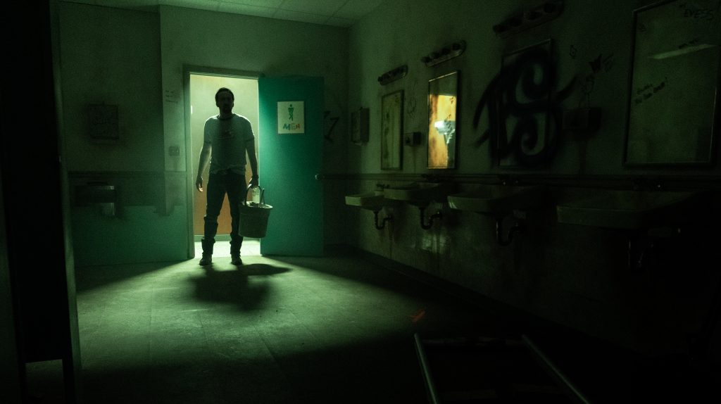 Nicolas Cage as the Janitor entering a dark and scary bathroom as seen in Willy's Wonderland directed by Kevin Lewis.
