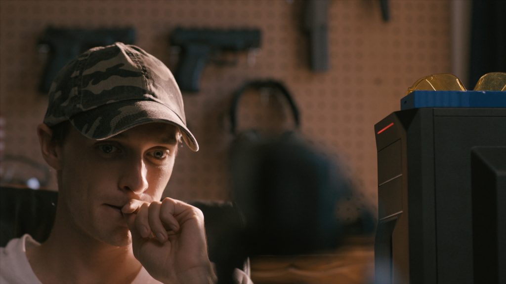 Will Madden as Adam sitting as his computer in front of a wall of guns as seen in the new film Beast Beast.