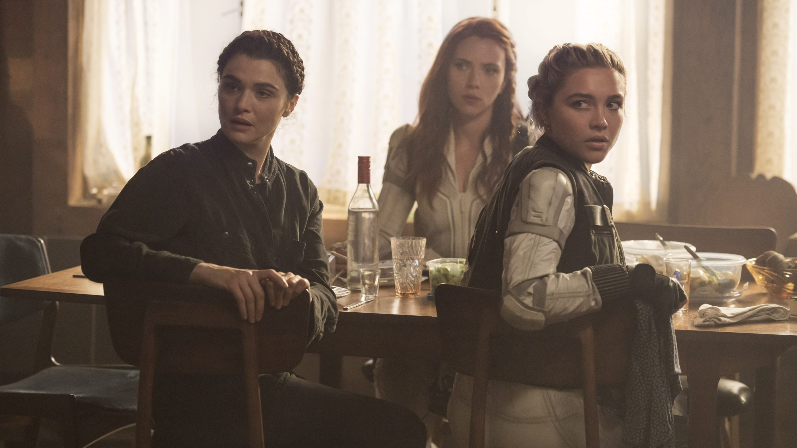 Rachel Weisz, Scarlett Johansson, & Florence Pugh sit at a dinner table while wearing spy costumes as seen in BLACK WIDOW, the latest MCU film to hit theaters.