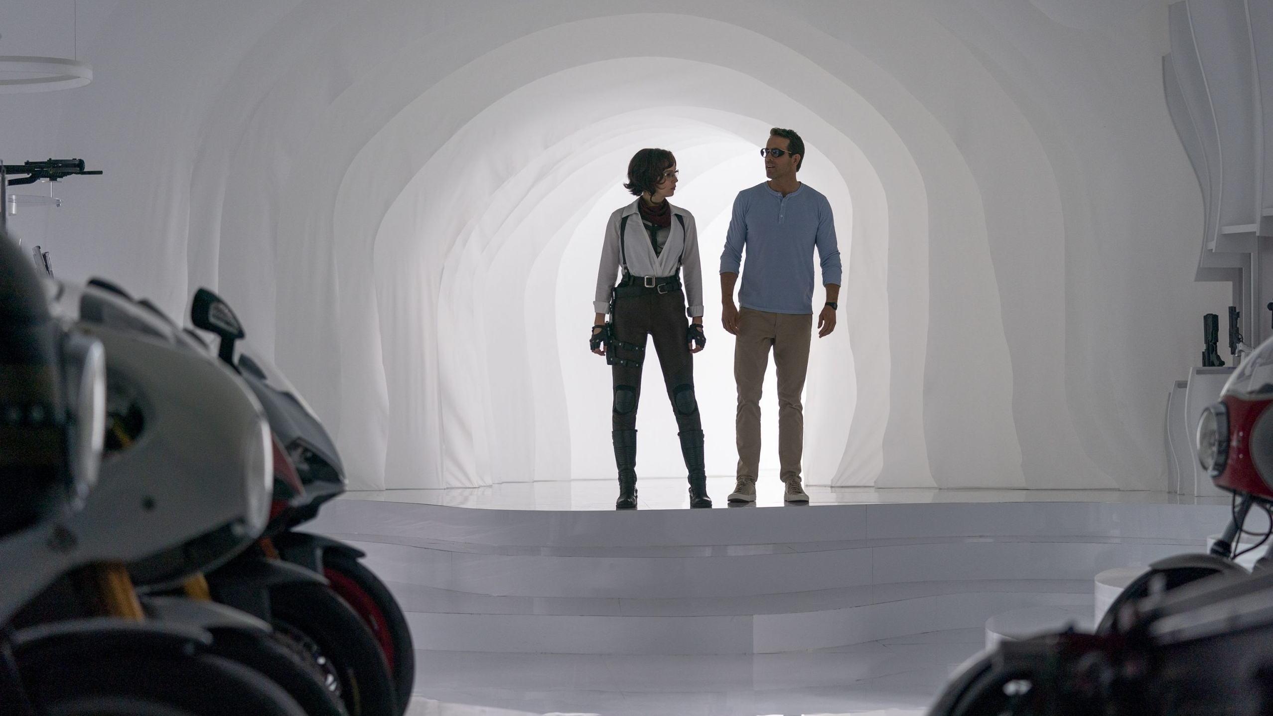 Jodie Comer as Molotov Girl and Ryan Reynolds as Guy enter a room filled with weapons and motorcycles through a white portal as seen in FREE GUY written by Matt Lieberman. 