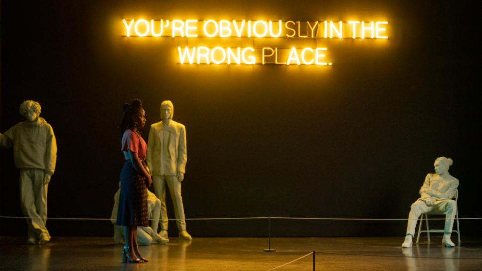Teyonah Parris standing in an art installation with white statues of men around her with a golden neon sign on the wall that reads "you're obviously in the wrong place" as seen in the new CANDYMAN.