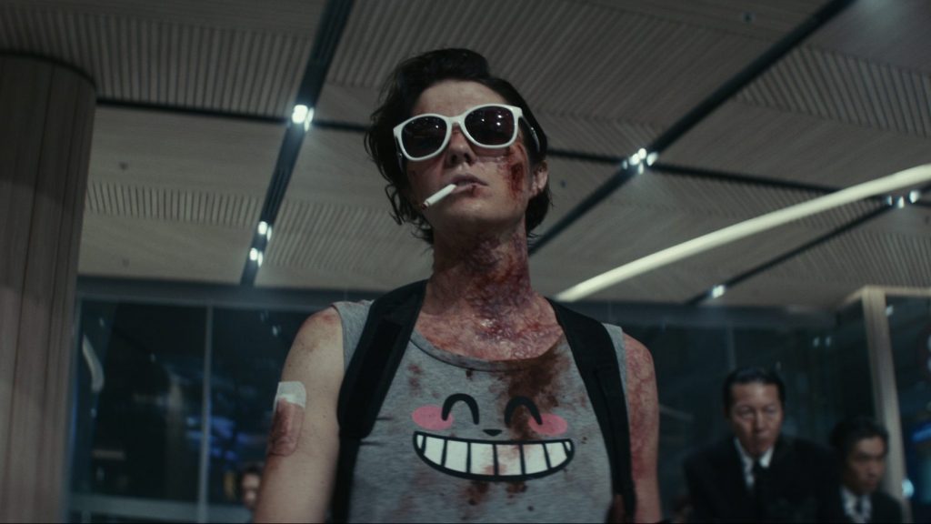 Mary Elizabeth Winstead looking nonchalant wearing big white sunglasses while smoking a cigarette as she is visibly ill with poising running through her veins, as seen in KATE coming to Netflix September 2021.