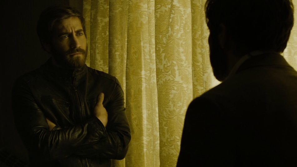 Two Jake Gyllenhaal doppelgangers stare at each other next to a golden yellow curtain inside a dark room as seen in ENEMY directed by Denis Villeneuve.