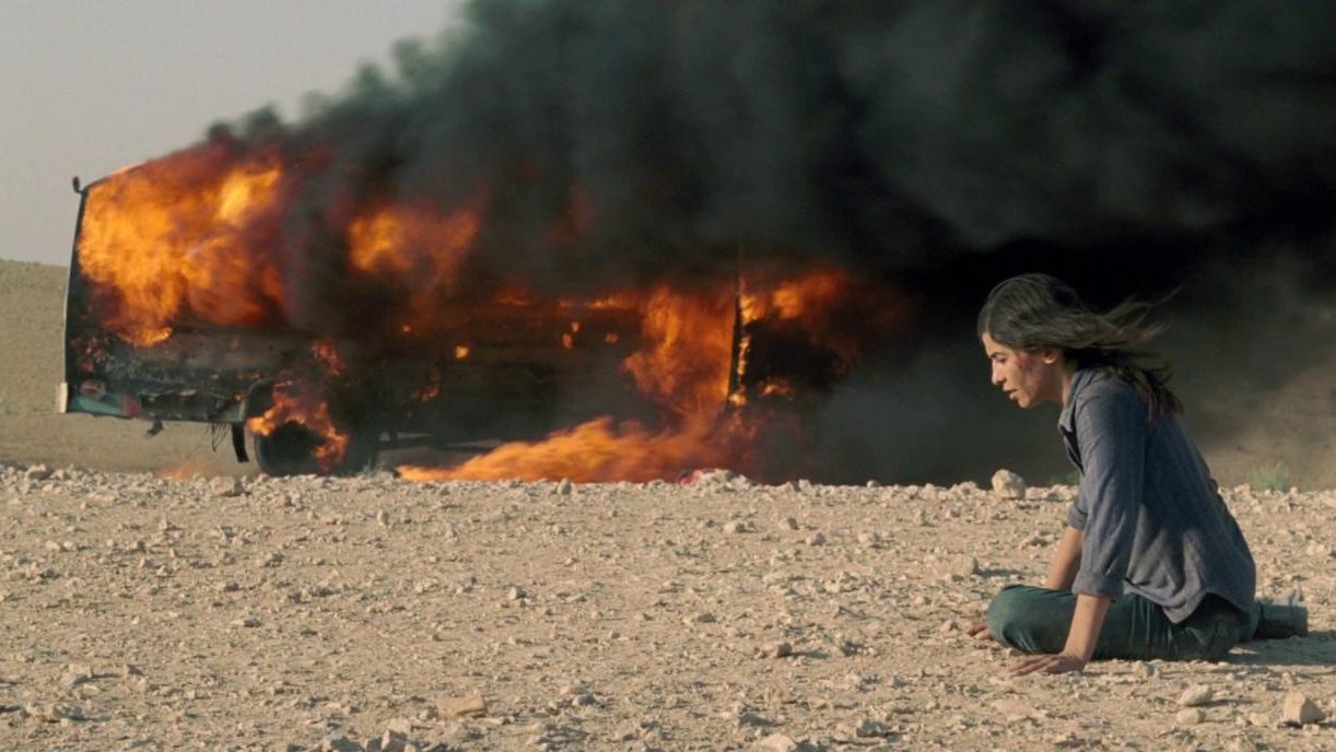 A woman lays on her needs with blood coming down her face in front of a truck engulfed in flames in the desert as seen in INCENDIES directed by Denis Villeneuve. 