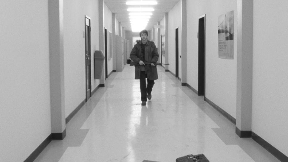 A black and white still of a young male serial killer holding an assault rifle walking down an empty school hallway from POLYTECHNIQUE directed by Denis Villeneuve. 