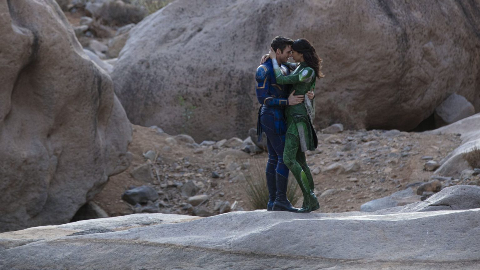 Richard Madden as Ikaris and Gemma Chan as Sersi embrace each other passionately while wearing their trademark blue and green cosmic suits in front of a rock formation as seen in the new Marvel Studios film ETERNALS directed by Chloé Zhao.