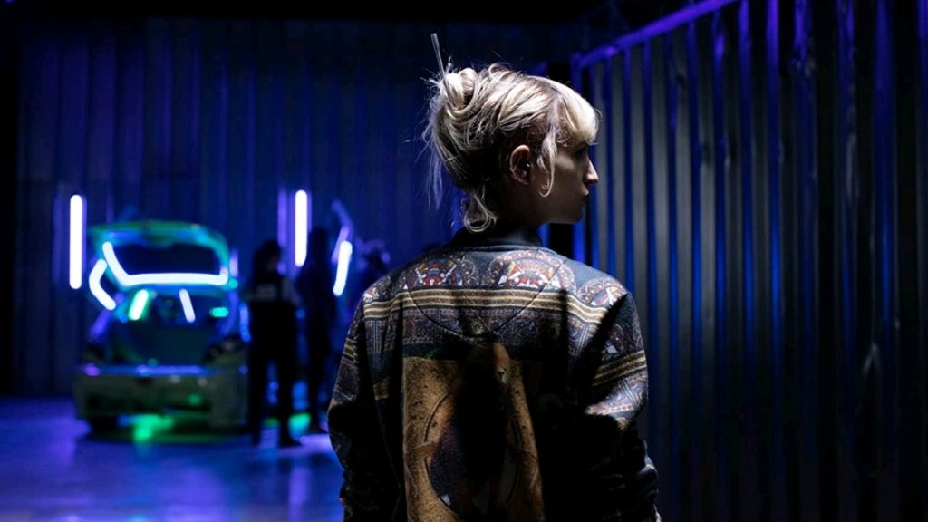 Agatha Rousselle as Alexia walking into a neon-lit underground car show as seen in TITANE directed by Julia Ducournau.
