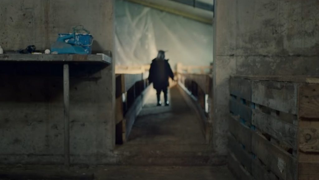 The half-human, half-lamb hybrid child Ada is obscured as she stands between some sheep pens in a barn as seen in the new A24 film LAMB. 
