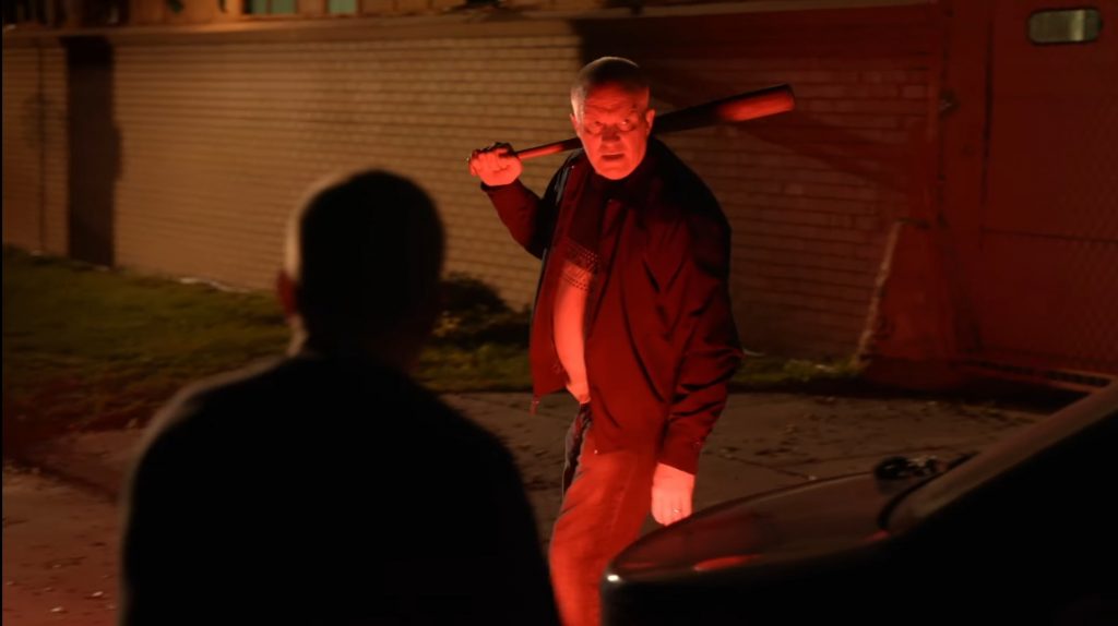 Anthony Michael Hall as Tommy Doyle paces around a car in bright red lights while holding a baseball bat over his shoulders as seen in HALLOWEEN KILLS directed by David Gordon Green. 