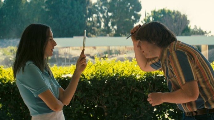 Alana Haim lifts a small mirror to Cooper Hoffman's face in annoyance as he combs his hair for picture day as seen in LICORICE PIZZA written and directed by Paul Thomas Anderson.