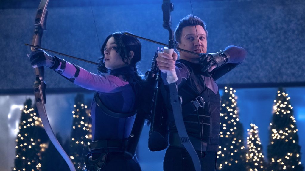 Hailee Steinfeld as Kate Bishop and Jeremy Renner as Clint Barton aim their bow and arrows side to side in front of some Christmas Trees as seen in HAWKEYE on Disney+ produced by Marvel Studios president Kevin Feige.