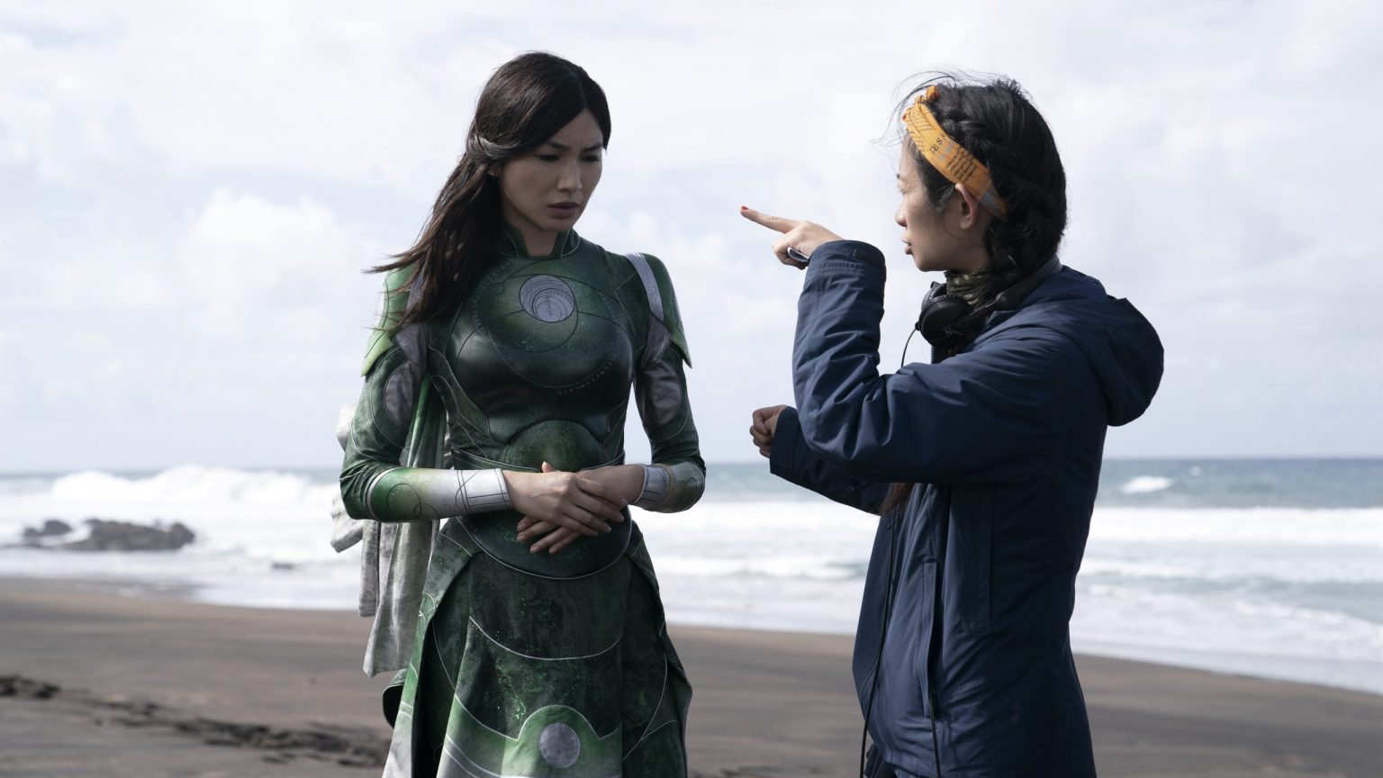 Oscar-winning director Chloé Zhao gives direction to Gemma Chan in her green costume as Sersi on a vast beach while shooting the new Marvel Studios film ETERNALS.