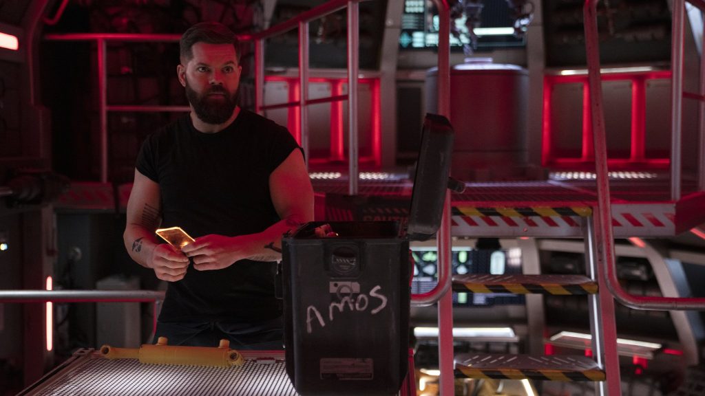 Wes Chatham as Amos Burton collecting supplies out of a chest labeled with his name on the deck of a space ship as seen in season 6 of THE EXPANSE on Amazon Prime Video. 
