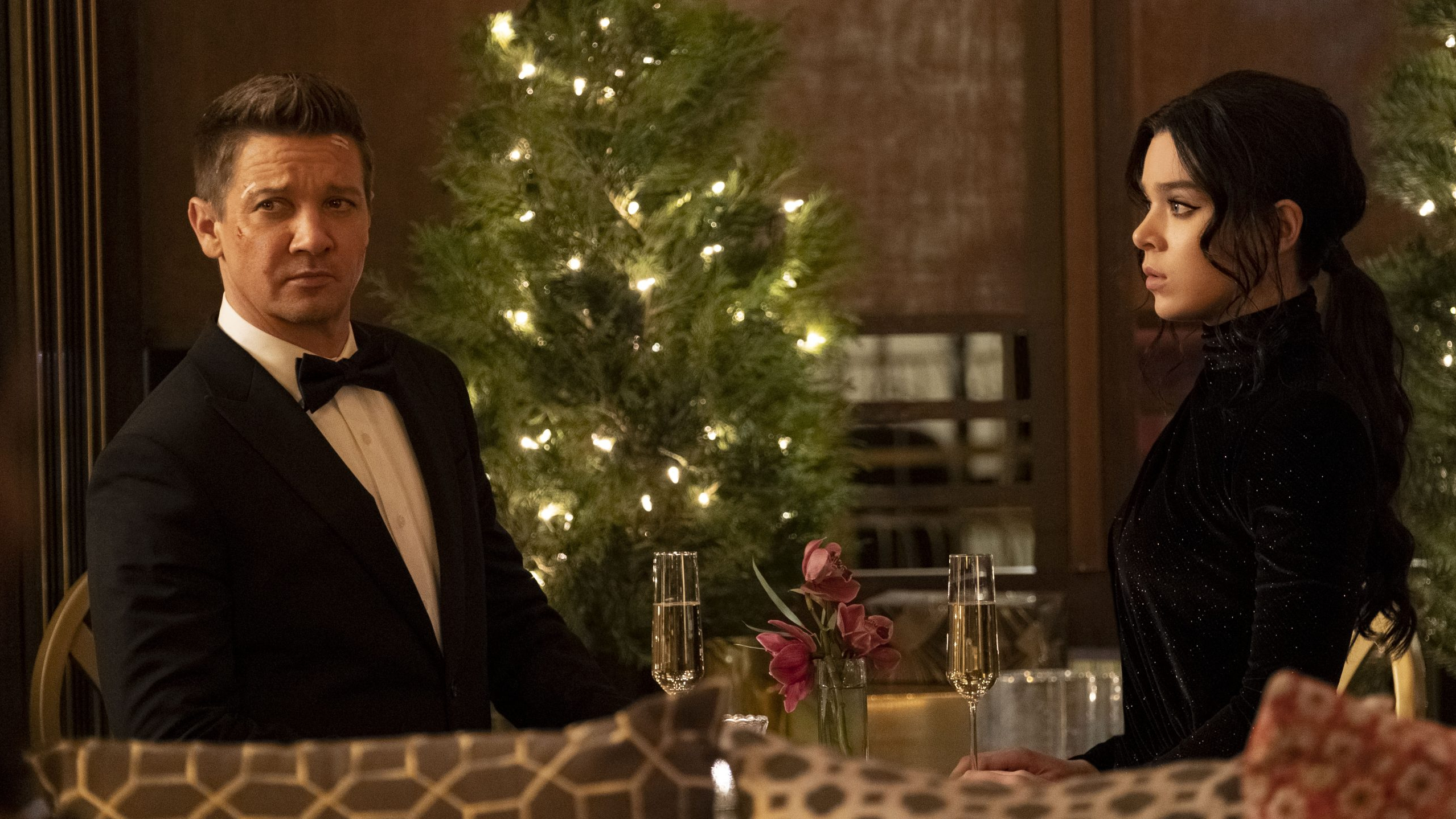 Jeremy Renner as Clint Barton in a tuxedo and bow tie shares champagne with Hailee Steinfeld as Kate Bishop in a black dress by a Christmas tree in the season finale of HAWKEYE on Disney+