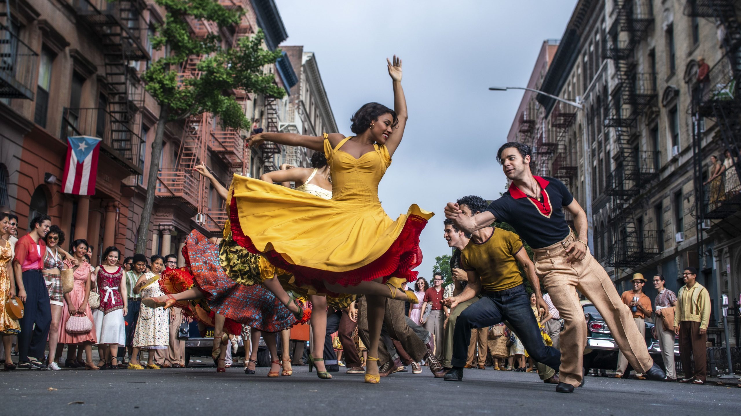 Ariana DeBose as Anita and David Alvarez as Bernardo dance in the middle of a lively crowd in the daytime New York streets in WEST SIDE STORY directed by Steven Spielberg, which features no subtitles for scenes in Spanish.
