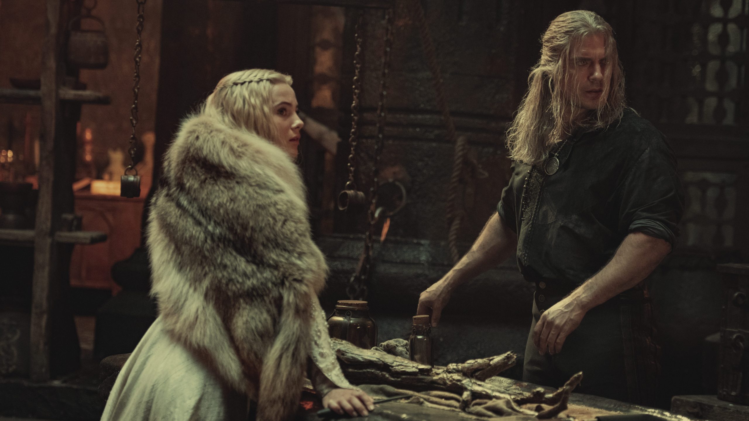 Freya Allan as Ciri wearing a white fur coat and Henry Cavill as Geralt stand together in shock as seen in season 2 of THE WITCHER on Netflix.