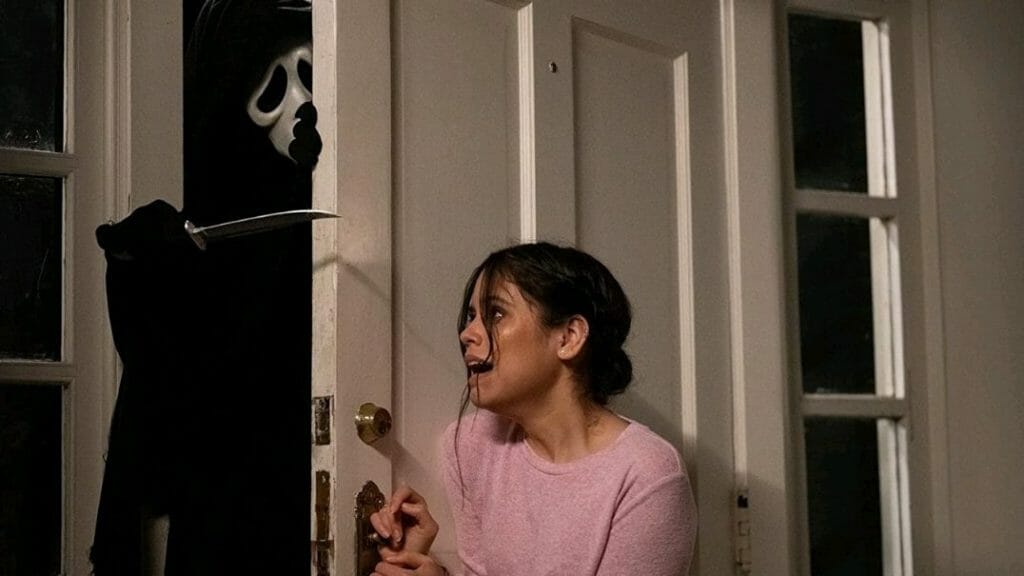 Jenna Ortega frantically holds the Ghostface killer back from behind a door as he tries to stick his arm inside and stab her with a sharp blade in SCREAM (2022).