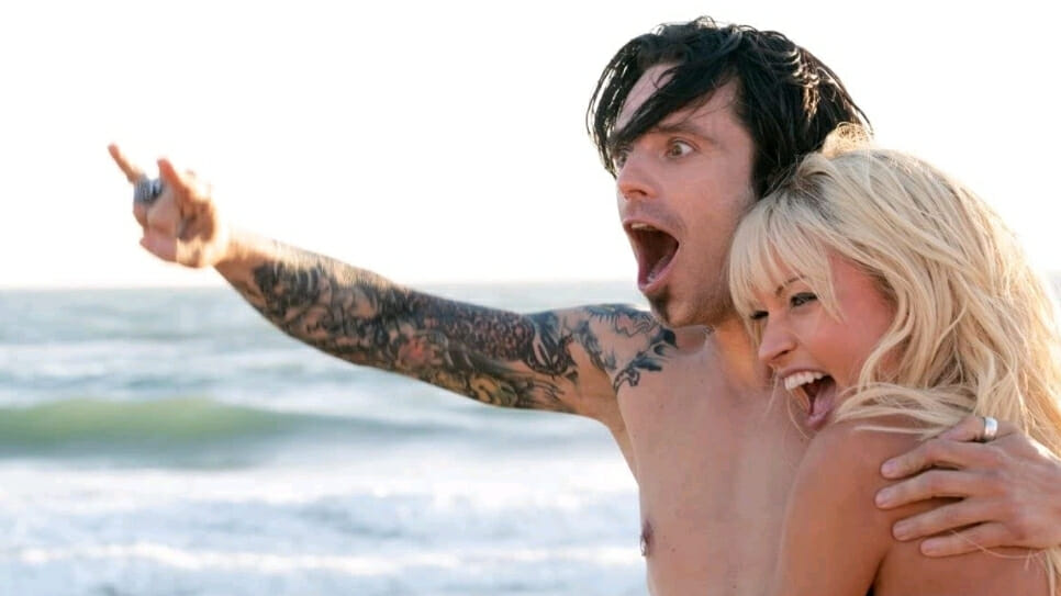 Sebastian Stan as Tommy Lee makes devil horns with one hand while holding Lily James as Pamela Anderson with the other as waves crash behind them on the beach as seen in the new Hulu miniseries PAM & TOMMY.