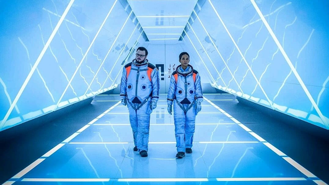 John Bradley and Halle Berry walk down a triangular chute with neon lights  in astronaut gear in MOONFALL directed by Roland Emmerich. 