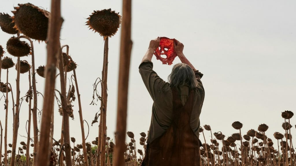 Leatherface holds up the bloody skinned face of one of his victims to use as his new mask while standing in a field of dried sunflowers as seen in TEXAS CHAINSAW MASSACRE on Netflix.