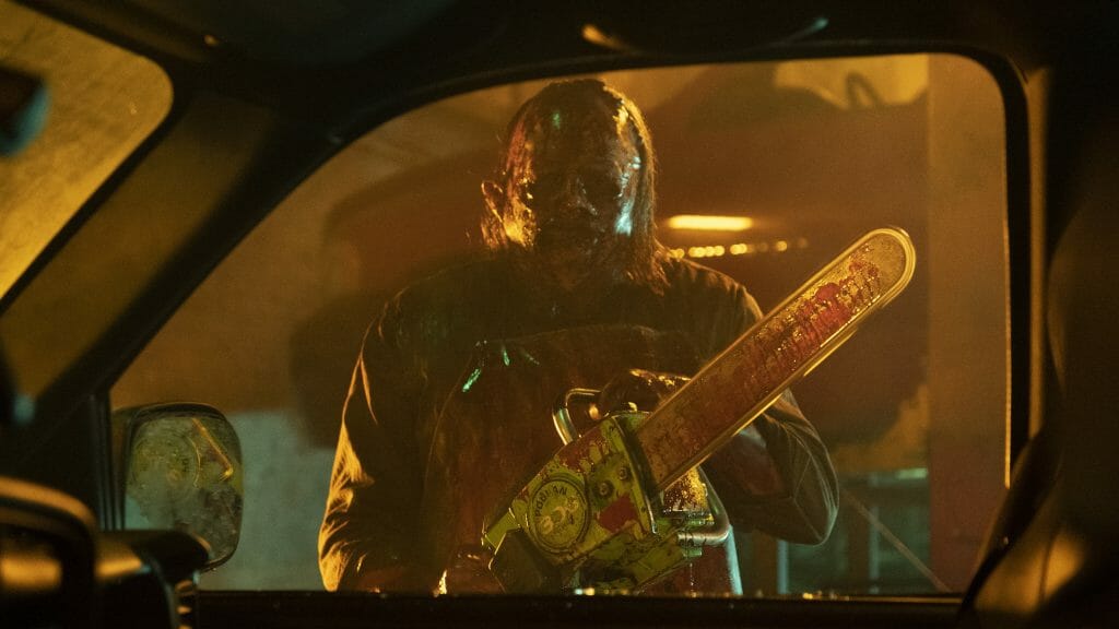 Leatherface covered in blood and holding a razor sharp chainsaw stares at his next victim from outside a car as seen in TEXAS CHAINSAW MASSACRE on Netflix. 