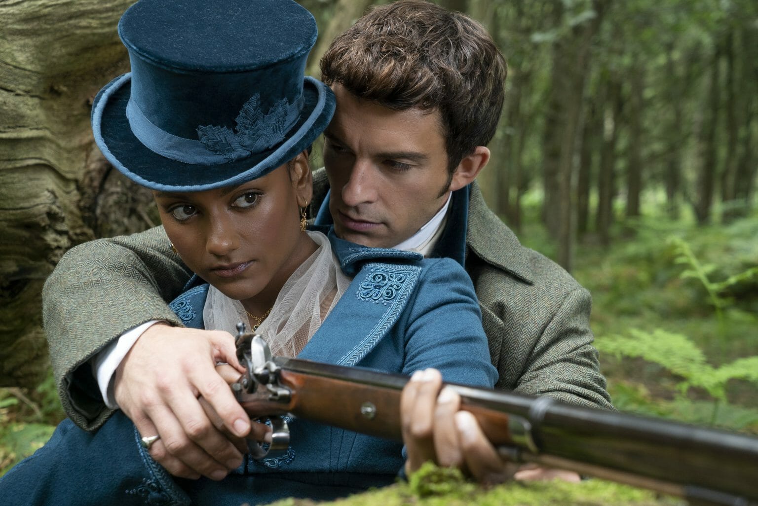Anthony Bridgeton played by Jonathan Bailey wraps his arms around Kate Sharma played by Simone Ashley as he teaches her how to shoot a rifle while they both wear fancy dress attire in season 2 of BRIDGERTON on Netflix.