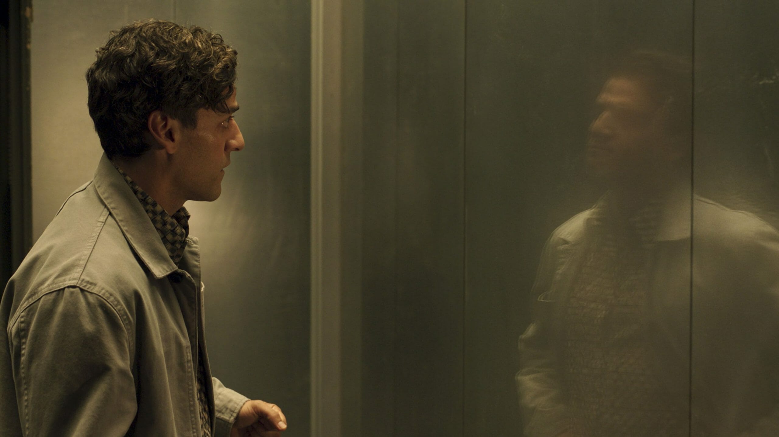 Oscar Isaac as Steven Grant stares at his reflection of his other identity Marc Spector on a shiny wall in the new Marvel Disney+ series MOON KNIGHT.  