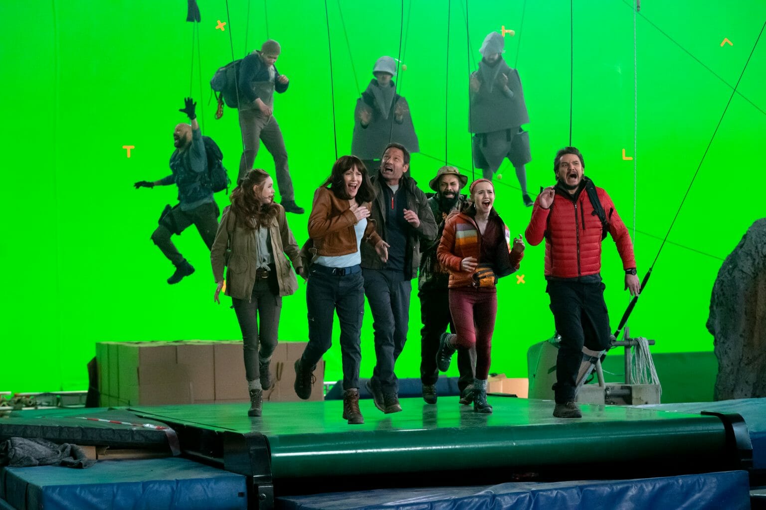 The main cast of THE BUBBLE pretend to run in fear on a large treadmill in front of a giant green screen while actors in digital reference dinosaur suits chase them on wires as seen in the new Netflix film directed by Judd Apatow.