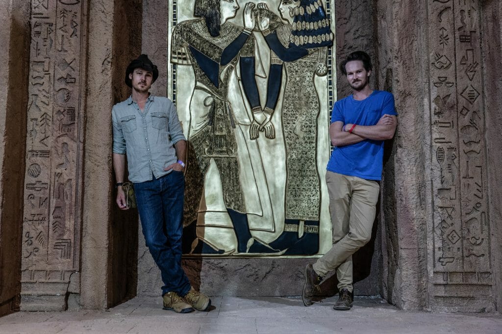 The Nee Brothers, Aaron Nee and Adam Nee, pose together and lean their backs on ancient temple walls with hieroglyphics on the set of the action rom-com THE LOST CITY.