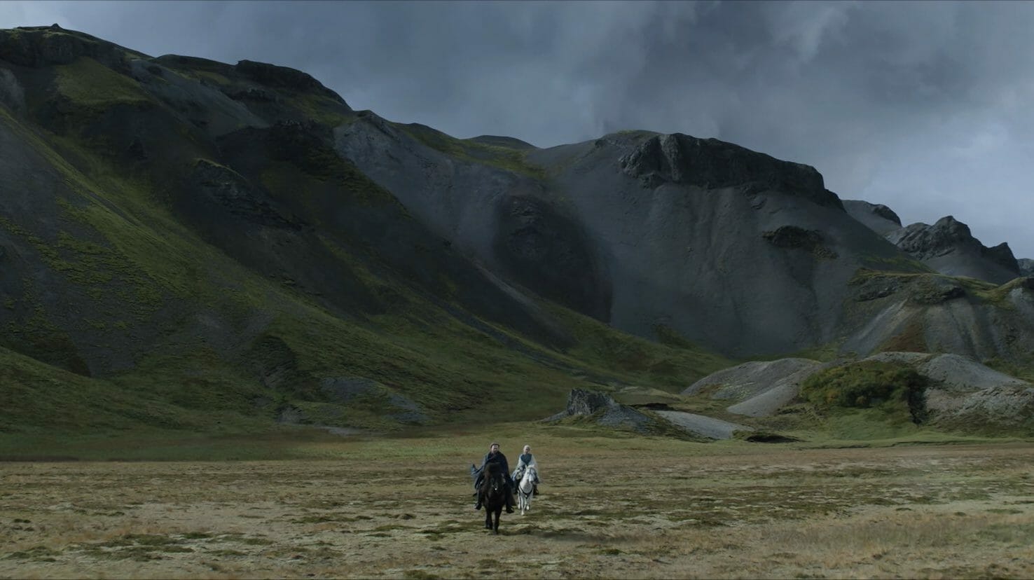 Alexander Skarsgård and Anya Taylor-Joy ride black and white stallions in front of dark charcoal mountains on Iceland's wide open grassy fields in THE NORTHMAN.  