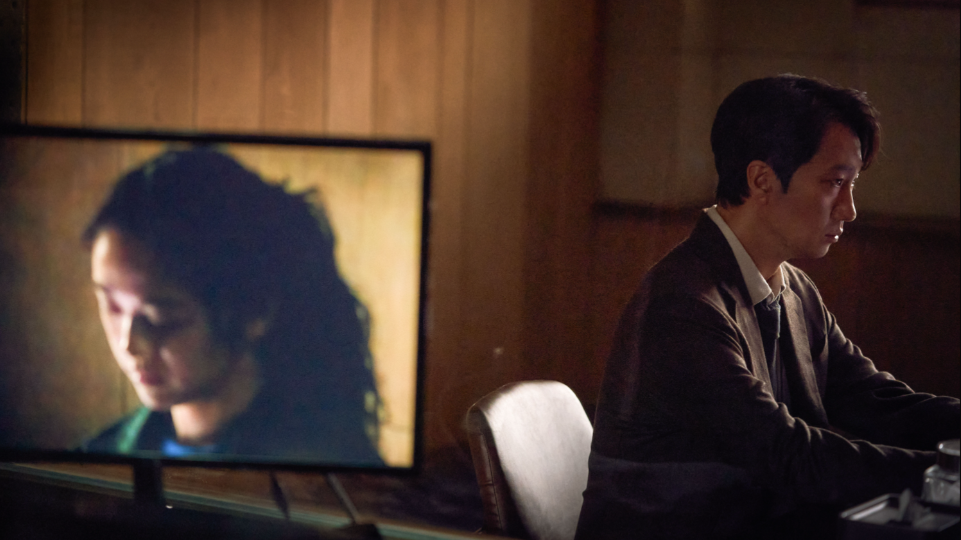 Park Hae-il as an insomniac homicide detective interrogates a new widower played by Tang Wei while her testimony is shown on a TV screen in DECISION TO LEAVE. 