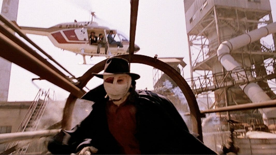 Darkman played by Liam Neeson runs away from a helicopter filled with mob goons shooting at him while on top of an industrial factory in DARKMAN directed by Sam Raimi. 
