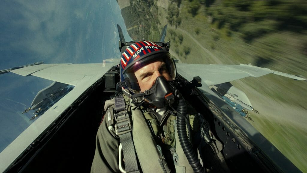 Tom Cruise as Captain Pete "Maverick" Mitchell makes a sharp left turn at high speed in his jet fighter while flying super low over a green valley in TOP GUN: MAVERICK.