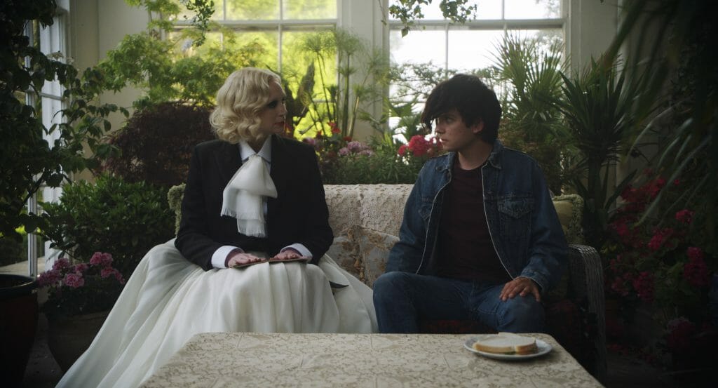 Gwendoline Christie wearing a flagrant black and white dress sits with Asa Butterfield wearing a jean jacket and jean pants awkwardly in a garden for lunch time in FLUX GOURMET.