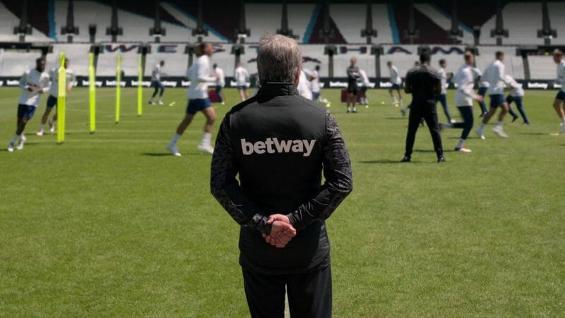 Nate played by Nick Mohammed stands in a villainous pose with his arms behind his back as he coaches a new team on the West Ham United soccer field from the TED LASSO Season 2 finale.  