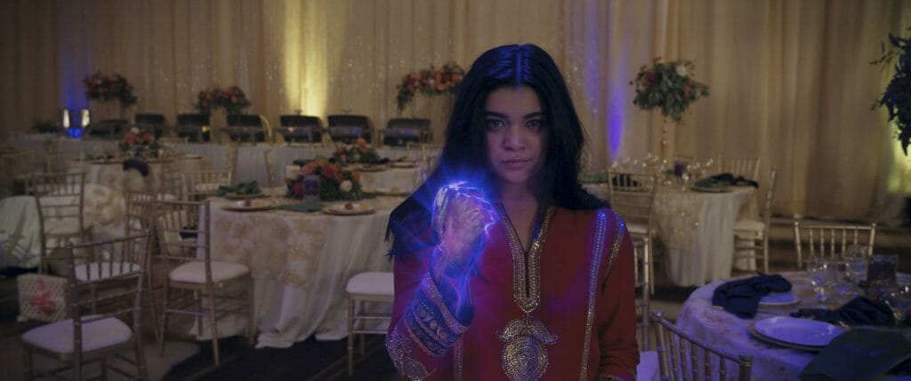 Iman Vellani as Kamala Khan strikes a battle pose with her purple glowing first in traditional celebratory Muslin attire at a family party in the MCU Disney+ series MS. MARVEL.