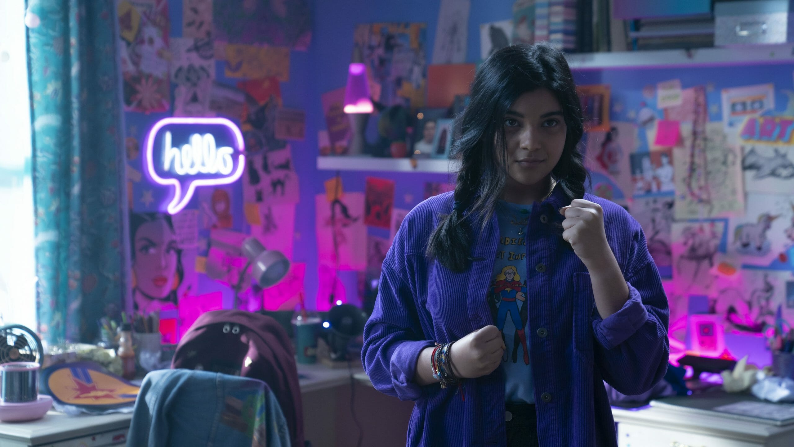 Iman Vellani as Kamala Khan strikes a battle pose with her fists while wearing a Captain Marvel shirt in her neon purple room filled with MCU superhero decorations in MS. MARVEL on Disney+. 