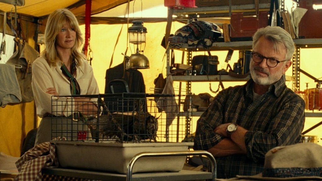 Laura Dern and Sam Neill examine a prehistoric locust trapped in a small cage from within an archaeologist tent filled with excavating tools in JURASSIC WORLD DOMINION. 