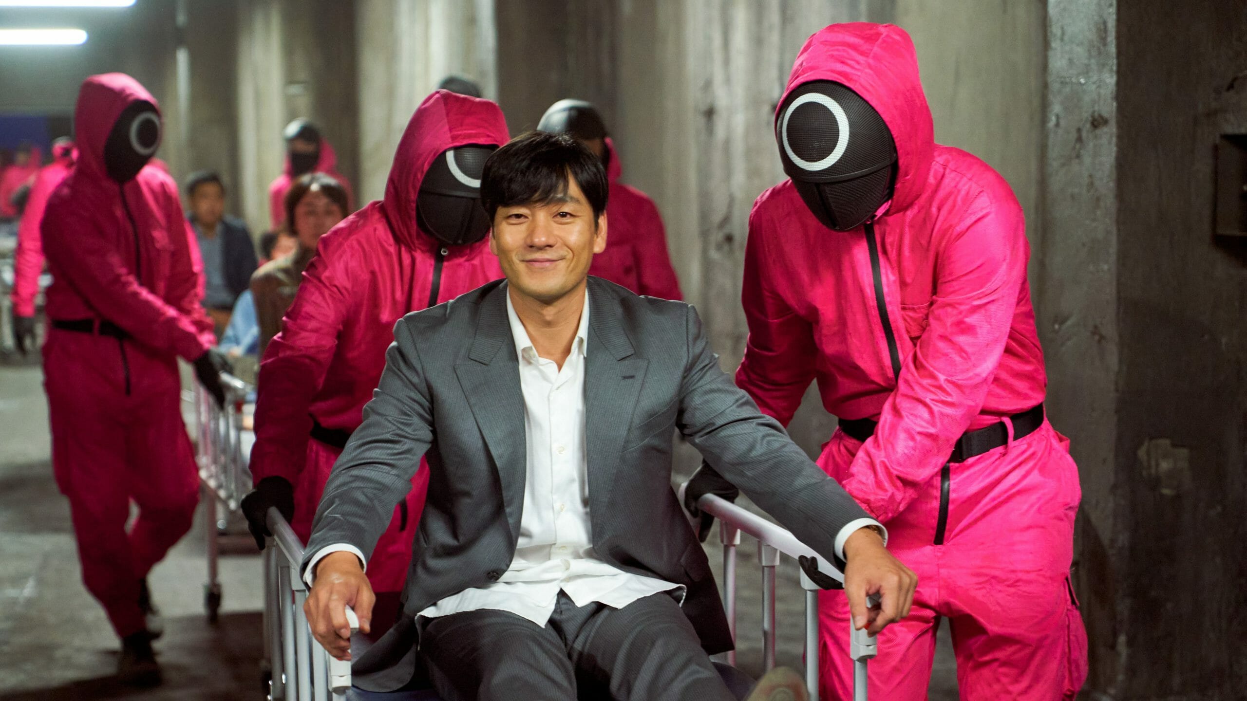 Park Hae-soo jokingly smiles as he's being pushed on a gurney by Squid Game guards wearing pink suits and black masks with white circles behind the scenes of the major Netflix series. 