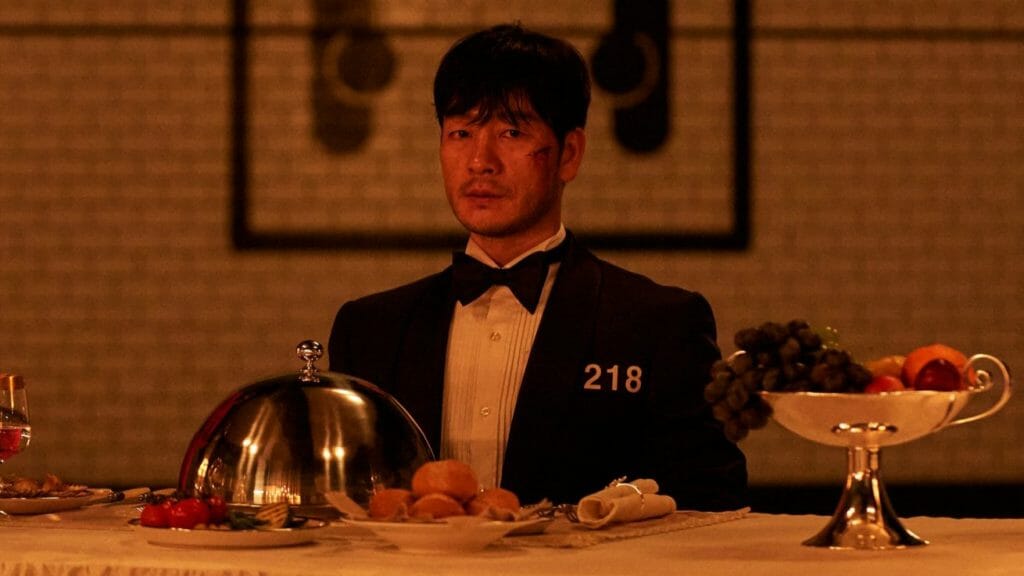 Park Hae-soo stares directly at the camera as he sits in cold sweat at a luxury banquet the night before the final round of Squid Game while wearing a tuxedo with his number 218 stitched on.