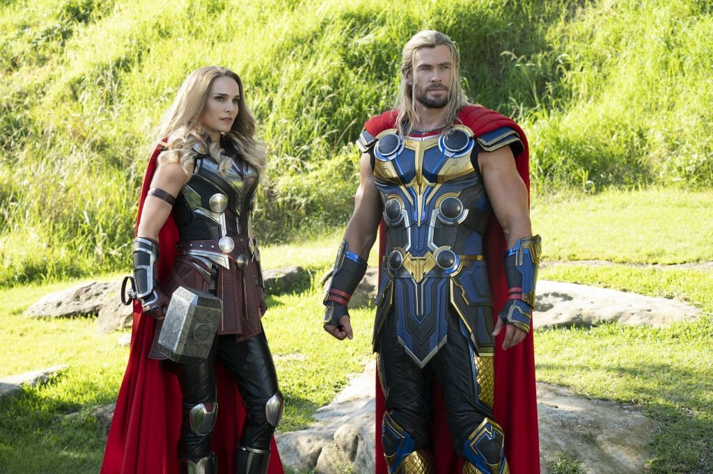 Natalie Portman as the Mighty Thor holding Mjölnir poses next to Chris Hemsworth as Thor Odinson holding Stormbreaker in THOR: LOVE AND THUNDER.