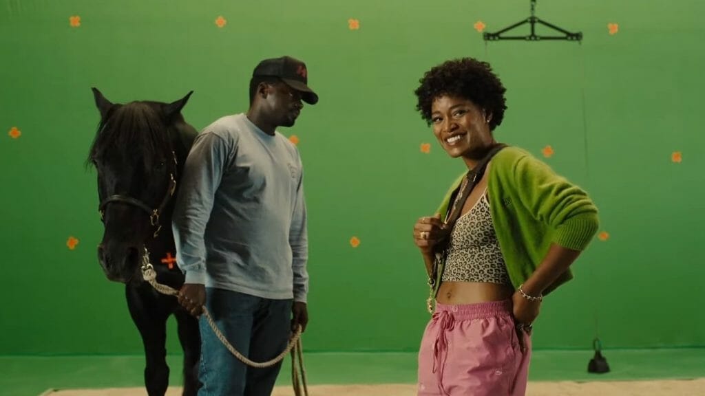 Daniel Kaluuya stands annoyed holding a horse in front of a green screen on a movie set while Keke Palmer gives an eccentric safety speech in NOPE directed by Jordan Peele.