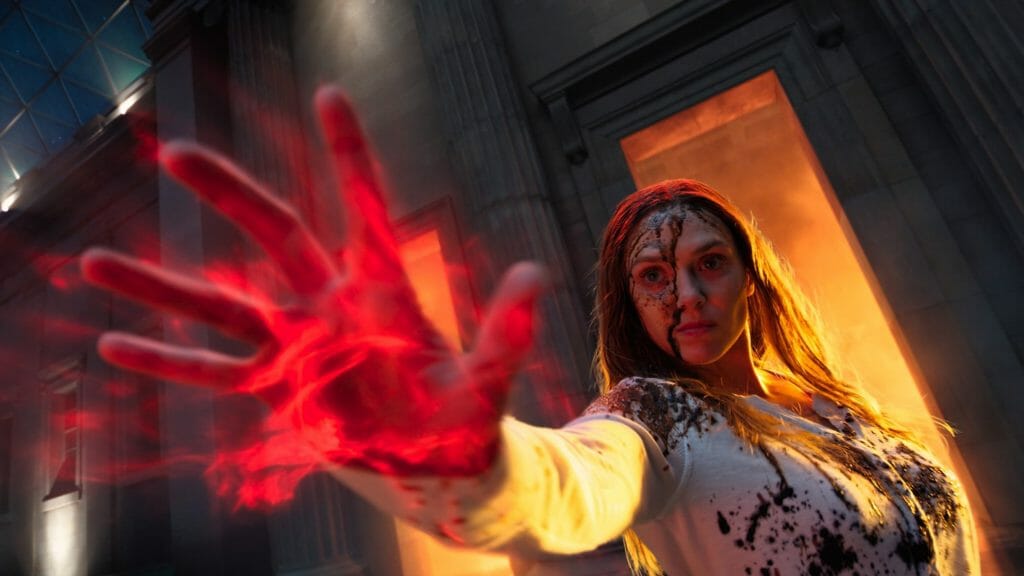 A close up of Elizabeth Olsen as the Scarlet Witch conjuring red magic energy from her hands as she prepares to attack in front of a burning fire in DOCTOR STRANGE IN THE MULTIVERSE OF MADNESS.