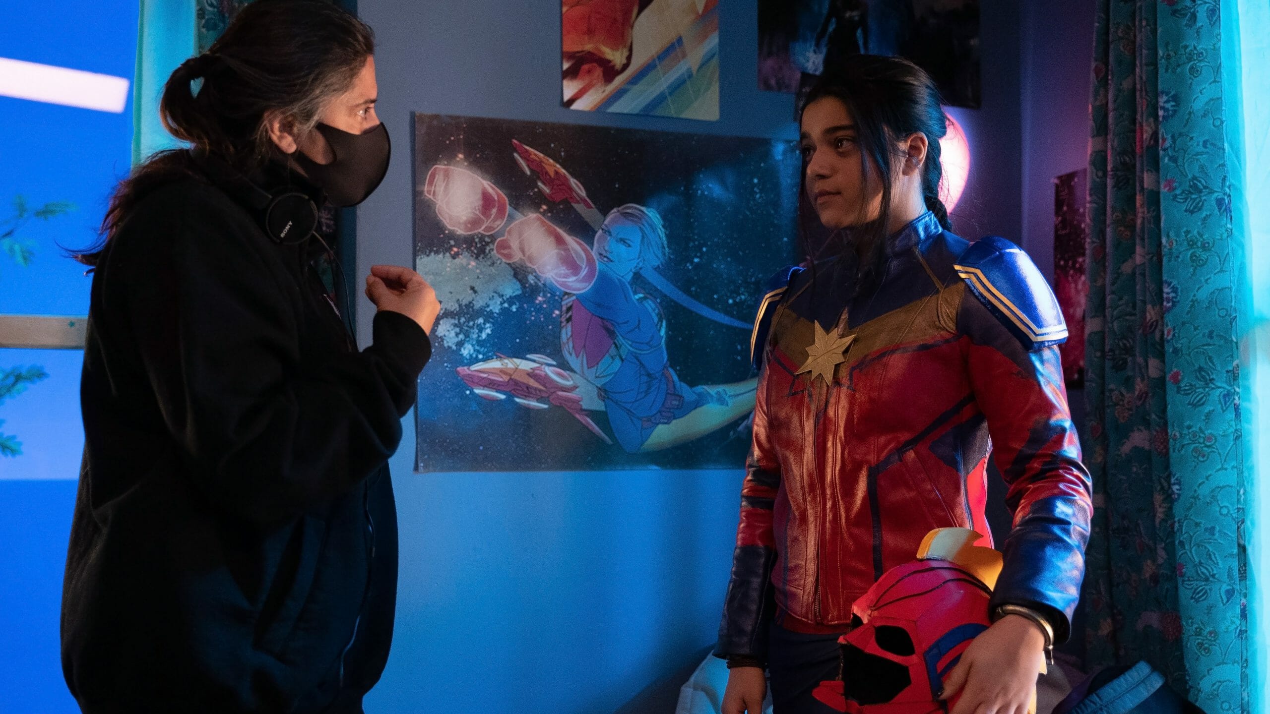 Director Sharmeen Obaid-Chinoy guides Iman Vellani in full Captain Marvel cosplay through a scene in her room full of superhero posters on the set of the MCU Disney+ series MS. MARVEL.
