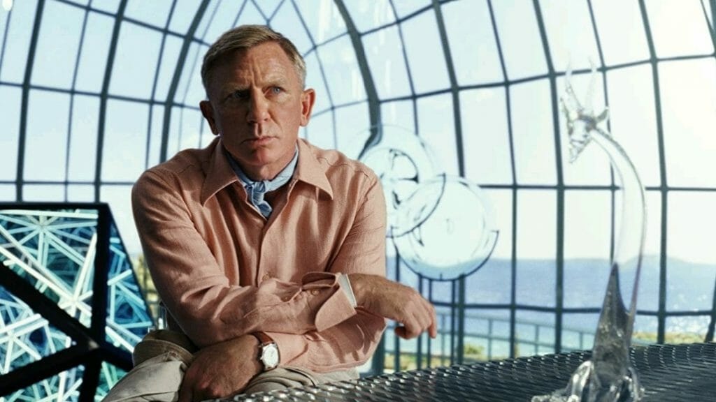 Daniel Craig as Benoit Blanc wearing a pink shirt and blue neck bandanna inside a giant glass dome building with an ocean view in GLASS ONION: A KNIVES OUT MYSTERY.